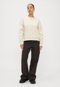 Κρεμ μπλούζα με στρογγυλή λαιμόκοψη, διακοσμημένη με διακριτικές λεπτομέρειες. Συνδυασμένη με σκούρες καφέ παντελόνες φαρδιάς γραμμής και μαύρα υποδήματα.