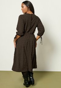 Eine Frau mit langen dunklen Haaren trägt ein strukturiertes, dunkelbraunes Midikleid mit 3/4-Ärmeln und schwarzen Ankle-Boots, während sie vor einem schlichten Hintergrund steht.