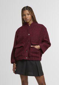 Giovane donna con i capelli intrecciati che indossa una giacca buttoned textured color bordeaux e una gonna plissettata nera, in piedi con una mano in tasca.