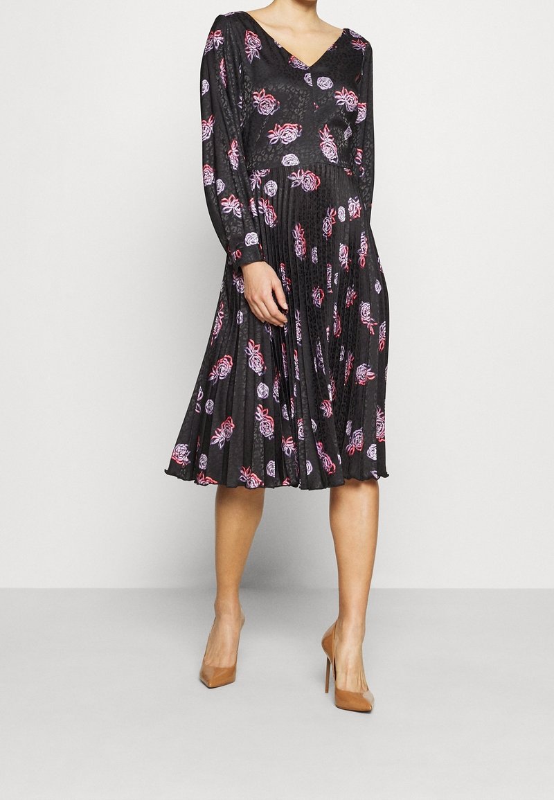 Robe noire à fleurs avec des manches longues, un décolleté en V et une jupe plissée. Des accents de fleurs roses et violettes ajoutent du détail à la texture du tissu.