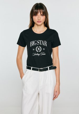 Kobieta ubrana w czarną koszulkę "BIG STAR Sailing Team" wpuszczoną do białych spodni z wysokim stanem i czarnym paskiem, stojąca na jednolitym tle.