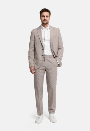 Man staat met één hand in zijn zak, draagt een beige pak, wit overhemd en witte sneakers tegen een effen lichte achtergrond.