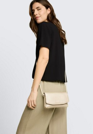 Vrouw met lang bruin haar, gekleed in een zwart kortärmelig shirt en beige broek, met een kleine beige crossbodytas over haar schouder.