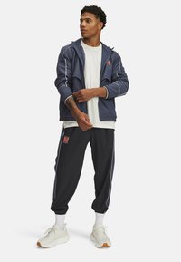 Marineblaues Jackett mit weißen Akzenten, kombiniert mit schwarzen Jogginghosen mit seitlichen Streifen und Logo. Darunter ein weißes T-Shirt und helle Sneakers.
