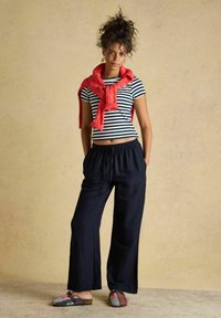 Woman with curly hair stands wearing black wide-leg pants, a striped cropped shirt, a red sweater tied around shoulders, and patterned slip-on shoes.