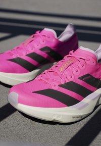 Pár jasnoružových bežeckých topánok Adidas Adizero s čiernymi pruhmi a bielymi podrážkami na šedom povrchu so tieňmi.