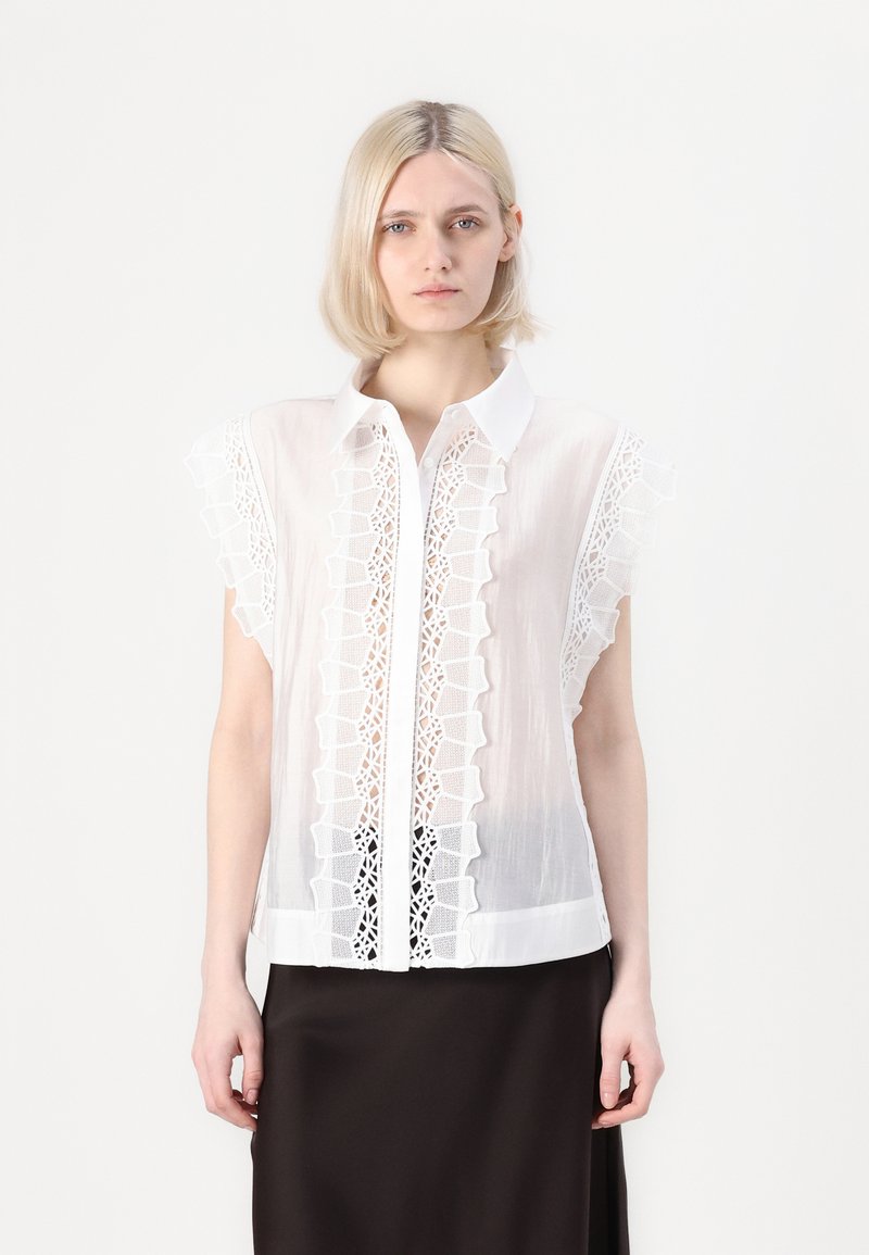Blusa bianca con maniche corte a volant e dettagli in pizzo ricamato sul davanti. Il tessuto è trasparente con chiusura a bottoni.