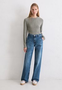 Vrouw met lang blond haar staat tegen een witte muur, gekleed in een grijze lange mouw top, high-waisted wijde blauwe jeans en witte sneakers.