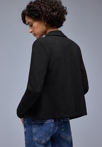 Femme aux cheveux bouclés et foncés portant une veste noire texturée et un jean bleu, légèrement tournée sur le côté, les mains dans les poches, devant un fond gris.