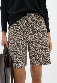 Femme portant un short à imprimé léopard et un pull noir à manches longues, tenant un sac cabas en cuir marron foncé.