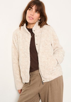 Frau trägt eine beige, flauschige Jacke mit Reißverschluss über einem braunen Pullover und braunen Hosen, steht mit einer Hand in der Tasche vor einer weißen Wand.