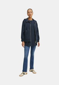 Felpa navy con zip e cappuccio regolabile, indossata sopra una tunica navy, abbinata a jeans a zampa di elefante blu chiaro e sneakers beige con accenti neri.
