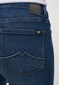 Tmavě modré džíny z denimu s výrazným vlnitým designem kapsy. Obsahují černou koženou visačku na pase a malou černou etiketu na kapse.