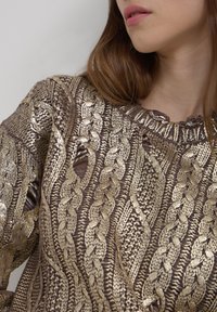 Pull en tricot avec un motif en câble doré métallique sur fond marron foncé. Tissu texturé avec un col rond et des découpes.