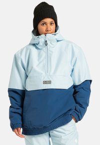 Jasnoniebieska i granatowa wiatrówka z zapięciem na zamek błyskawiczny przy szyi, elastycznymi mankietami oraz kieszenią z przodu; zawiera naszywkę z logo na klatce piersiowej.