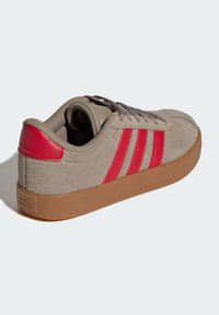 Bež teniska od antilop kože s gumenim potplatom, crvenom zakrpom na peti i tri crvene pruge sa strane, prikazana iz stražnjeg lijevog kuta.