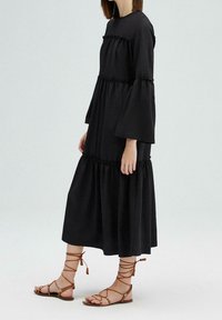 Robe noire à manches longues avec des volants en biais et un bas évasé. Accompagnée de sandales gladiateurs à lanières marron. Texture de tissu lisse.