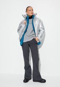 Vrouw in een zilveren pufferjack met blauwe fleece voering, grijze sweatshirt, zwarte broek en zwarte schoenen, staande tegen een witte muur.