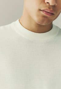 White knit sweater with a textured pattern, featuring a ribbed crew neckline. The fabric has a lightweight and breathable appearance.