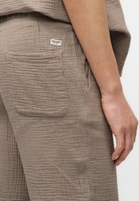 Κοντινή λήψη μπεζ υφασμάτινου παντελονιού με πίσω τσέπη και λευκή ετικέτα «JACK & JONES EST. 1990», δίπλα σε ένα χαλαρό χέρι που ακουμπά κοντά.