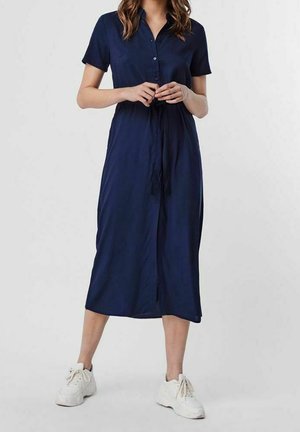 Mujer con un vestido midi azul marino de manga corta con botones, cinturón en la cintura y zapatillas blancas, de pie frente a un fondo sencillo.
