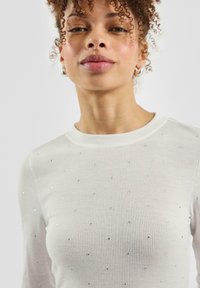 Weißes, geripptes Langarmshirt, besetzt mit kleinen, funkelnden Strasssteinen, die gleichmäßig über den Stoff verteilt sind. Klassischer Rundhalsausschnitt.