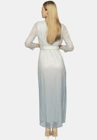 Srebrna długa sukienka o pionowych fałdach, z długimi rękawami, z wcięciem w talii i zwiewnym dołem, zestawiona z beżowymi szpilkami.