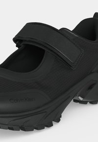 Черна маратонка Calvin Klein с текстуриран горен слой, дебела грапава подметка и широк регулируем лент с лепенка през горната част.