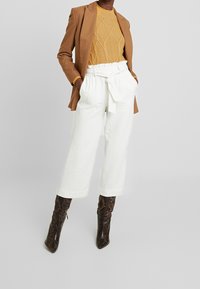 Blazer marron, pull en tricot moutarde texturé, pantalon blanc à jambes larges avec ceinture à nouer, et bottines marron à imprimé serpent.