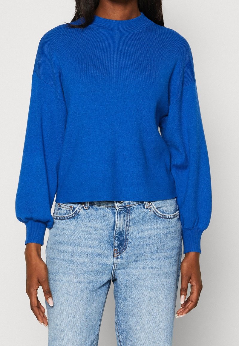 Femme portant un pull bleu vif à manches longues avec des poignets côtelés et un jean taille haute délavé clair sur un fond uni.