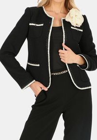 Czarna marynarka z tweedu z białymi dodatkami, z dwiema kieszeniami na piersiach, teksturowanym wykończeniem oraz kremowym kwiatem z materiału akcentującym ramię.