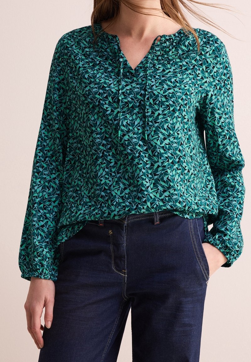 Femme portant un chemisier à manches longues avec motif bleu sarcelle et noir, col à cordon, et un jean bleu foncé, une main dans la poche.