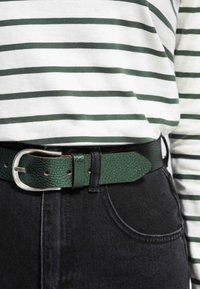 Grøn struktureret læderbælte med en sølvmetalspænde, båret over sorte jeans, parret med en hvid og grøn stribet skjorte.