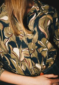 Blumig gemustertes Hemd mit dunkelblauem Grundton, olivgrünen und cremefarbenen Blumen, spitzem Kragen und durchgehender Knopfleiste, mit Gürtel gehalten.