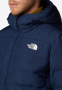 Granatowa kurtka puchowa z pełnym zamkiem błyskawicznym i wysokim kołnierzem; na lewej stronie znajduje się białe logo The North Face. Izolacyjny design.