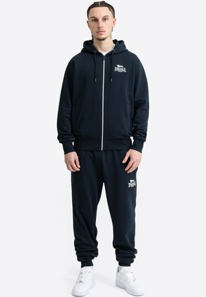 Man draagt een zwarte Lonsdale London hoodie en bijpassende joggers met witte sneakers, staand tegen een effen witte achtergrond.