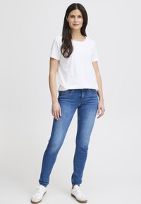 Balti medvilninė marškinėliai derinami su mėlynomis siaurėjančiomis džinsais. Modelis vilki baltus sportbačius su smėlio spalvos detalėmis, stovintis prieš paprastą foną.