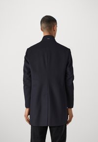 Cappotto di lana navy con colletto alto, taglio dritto e piega sul retro. Presenta un design minimale senza hardware o motivi visibili.
