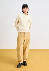 Sweatshirt crème, pantalon beige à jambe large et chaussures noires. Le sweatshirt présente une petite étiquette de logo ; le pantalon est en velours côtelé avec une finition texturée.
