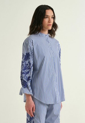 Donna con camicetta a righe blu e bianche con ricamo floreale sulle maniche e pantaloni a righe abbinati, in piedi di profilo.