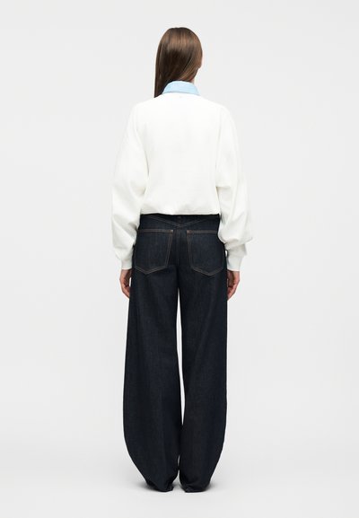 Γυναίκα με μακριά καστανά μαλλιά φοράει ένα λευκό μακρυμάνικο μπλουζάκι με μπλε γιακά και πλατύτερα σκουρόχρωμα τζιν παντελόνια, στέκεται με την πλάτη προς την κάμερα.