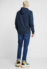 Sudadera con capucha de color azul marino, hecha de tejido suave, con un bolsillo canguro y puños acanalados, combinada con vaqueros azules y zapatillas multicolores.