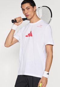 Valge spordisärk punase Adidas logoga, ümarsärg, lühikeste varrukatega, mustade lühikeste pükstega ja valge käepaelaga, tennisereketit hoides.