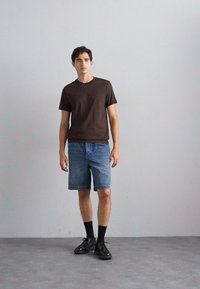 Camiseta de cuello redondo marrón, pantalones cortos de mezclilla, zapatos negros y calcetines negros, de pie frente a una pared y un suelo de color gris claro.
