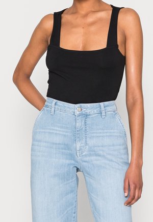 Vrouw draagt een zwart mouwloos topje met een vierkante hals, ingestopt in lichtblauwe high-waisted spijkerbroek, met één hand op de heup.