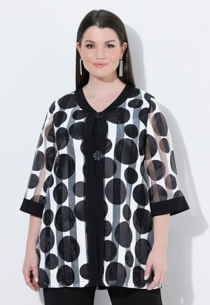 Femme portant une veste transparente noire et blanche avec de grands pois noirs et des rayures verticales, posant sur un fond blanc.