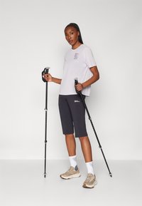 Lys lavendel kortermet skjorte med logo, grå knelange shorts og beige tursko. Svarte turstaver holdt i begge hender.