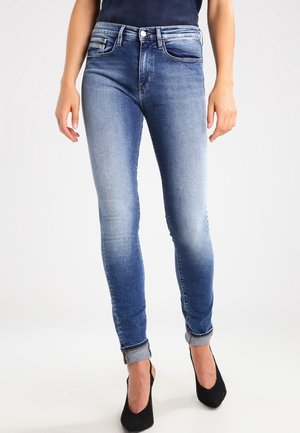 Enge blaue Jeans aus Denim mit verblasster Waschung, hoher Taille und umgeschlagenen Bündchen. Kombiniert mit schwarzen High-Heels.
