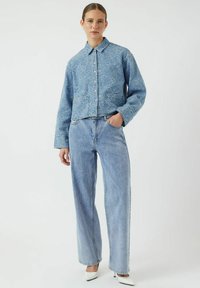 Giacca in denim azzurro chiaro con un motivo testurizzato, chiusura con bottoni, tasca sul petto, abbinata a jeans a gamba larga azzurro chiaro e tacchi bianchi.