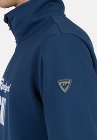 Giacca a pullover navy realizzata in materiale elastico, con colletto alto e una patch in gomma nera sullo spalla sinistra.